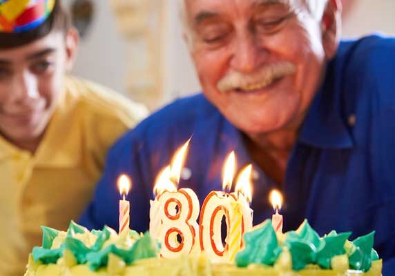 A smiling elderly man blows out birthday candles in the shape of the number 80