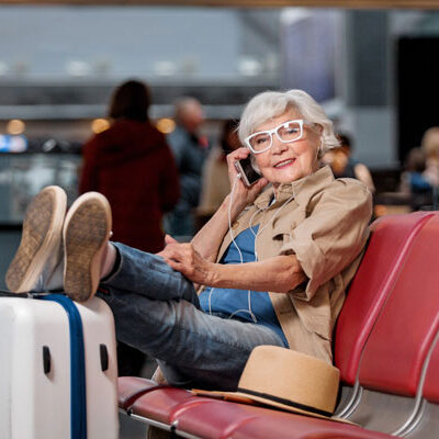 An elderly woman talks on a smartphone while sitting at an airport with her feet up on a suitcase